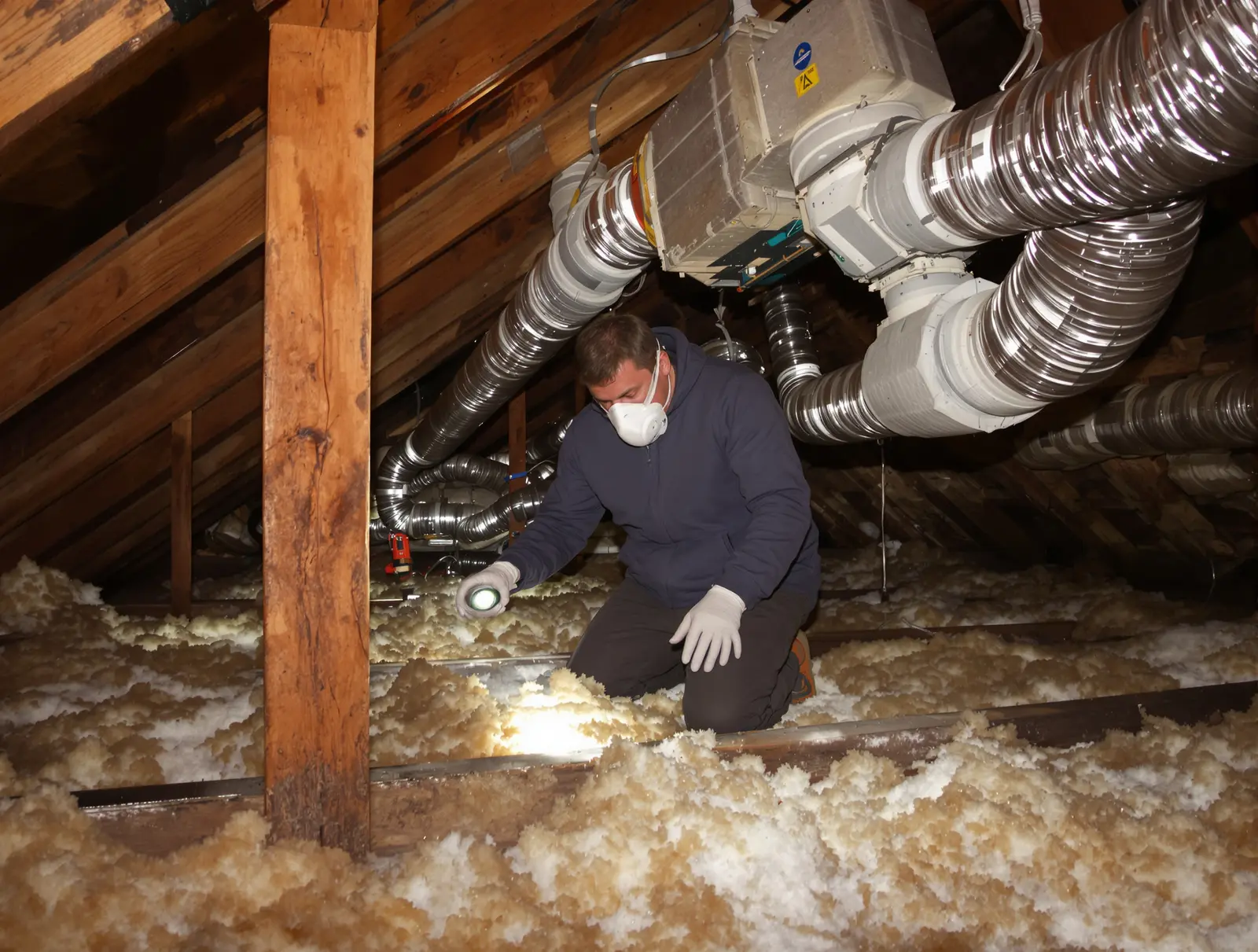 Person inspecting ductwork connections in an attic with a flashlight
